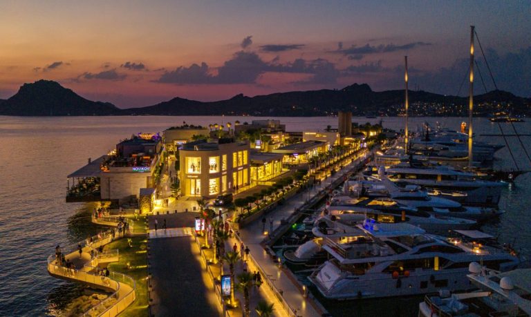 Aerial dusk view of superyachts docked at Yalıkavak Marina in Bodrum, Türkiye, with the marina's retail promenade lit up